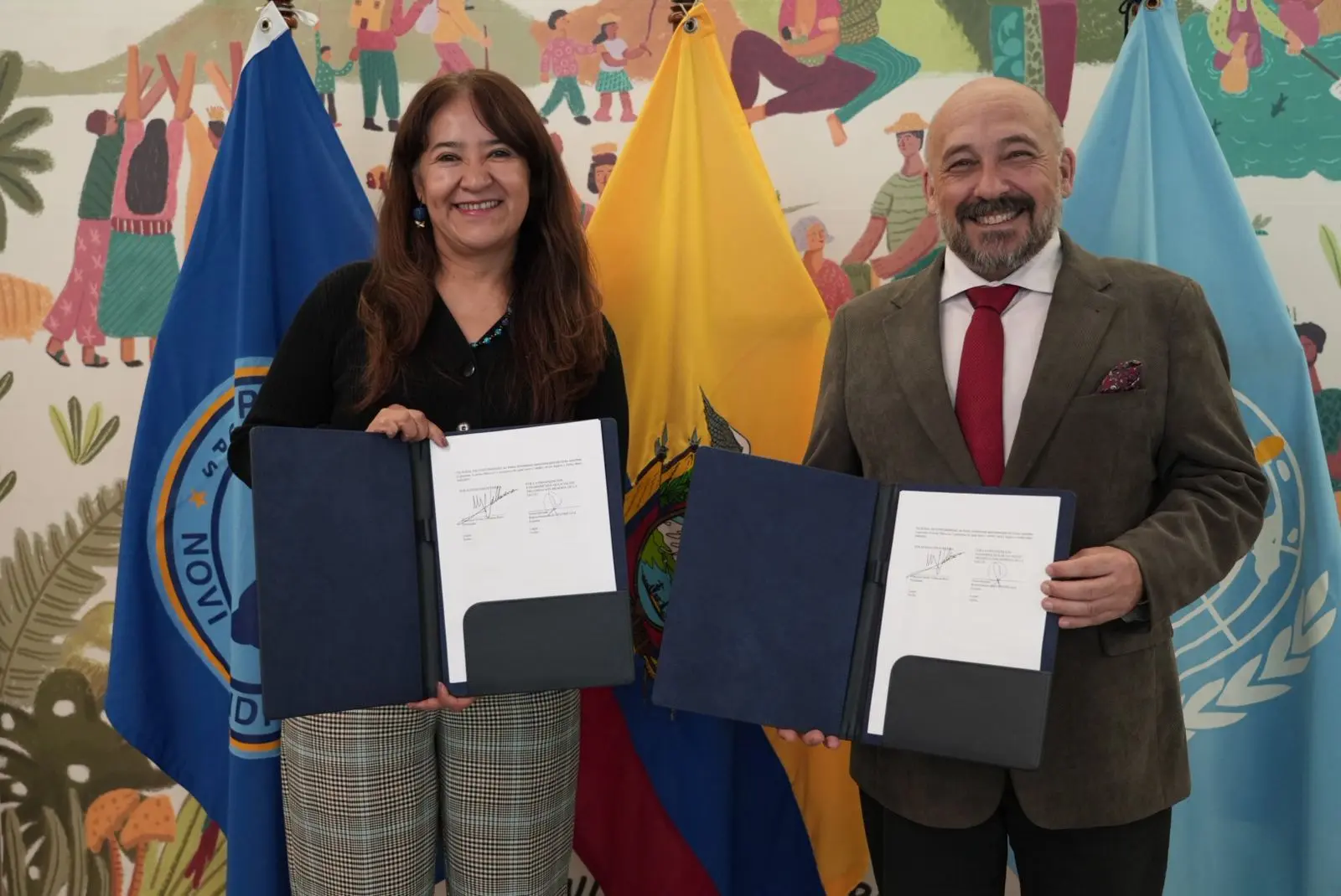 Un hombre y una mujer delante de un mural y unas bandeas enseñan unos papeles firmados